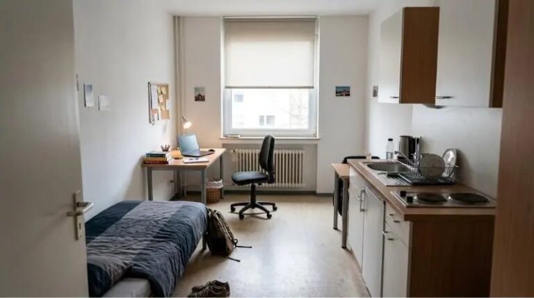Studentenapartment in einem Wohnheim: Einzelbett, Schreibtisch mit Laptop am Fenster, Pinnwand und schmale Küchenzeile mit zwei Kochfeldern.